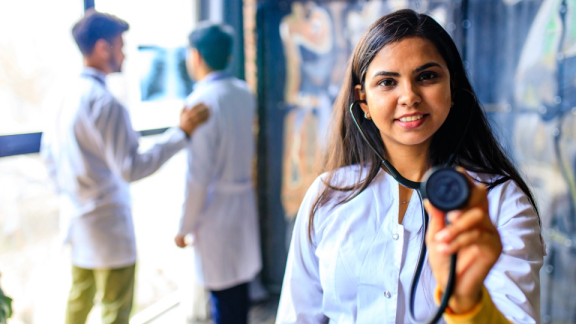 Eine marokkanische Ärztin hält ein Stethoskop zur Kamera, im Hintergrund besprechen sich zwei weitere Ärzte