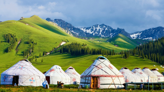 Traditionelle Jurten in grüner Berglandschaft der Mongolei mit schneebedeckten Gipfeln im Hintergrund