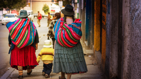 Zwei Bolivianerinnen in traditionellen Gewändern laufen mit einem Kind durch eine Straße
