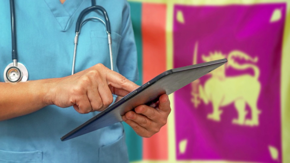 Ein Arzt aus Sri Lanka steht vor der Landesflagge und tippt etwas in ein Tablet