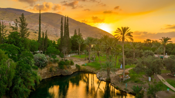 Ein Gewässer in einer trockenen Gegend in Israel mit Bäumen, Palmen, grünen Wiesen und Bergen im Hintergrund