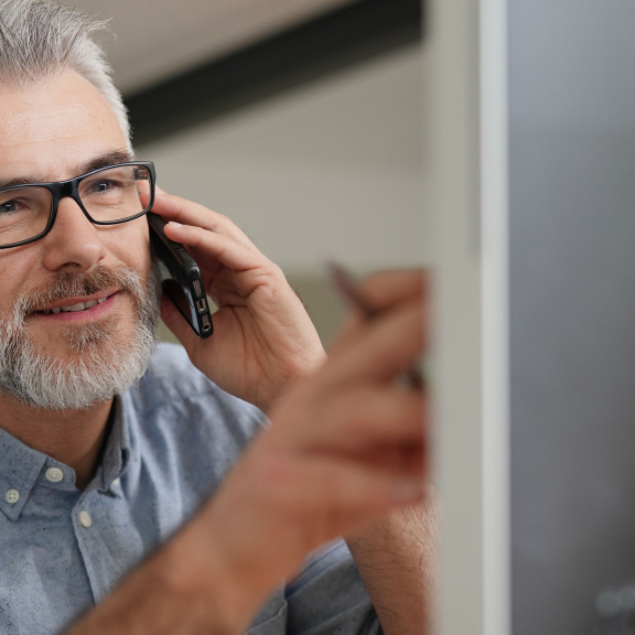 Freundlicher Mann mit Brille telefoniert am Computer, schaut sich eine FAQ-Frage an.