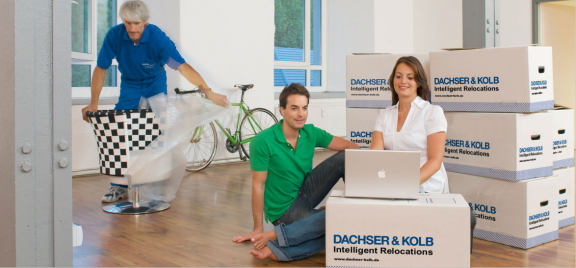 Paar zieht mit Umzugsservice in neues Zuhause um – Kisten, Laptop und Möbelpacker im modernen Wohnraum.