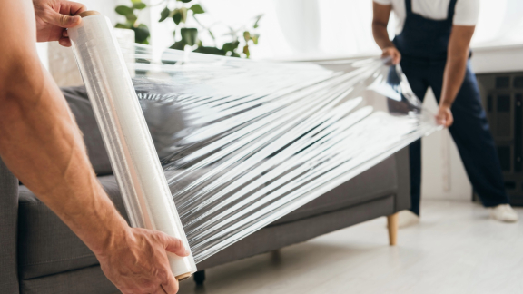 Two professional movers are wrapping a couch in transparent plastic wrap for a move.