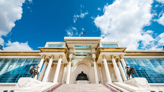Monumentales Regierungsgebäude in Ulaanbaatar mit Säulen, Treppe und mongolischer Flagge unter blauem Himmel