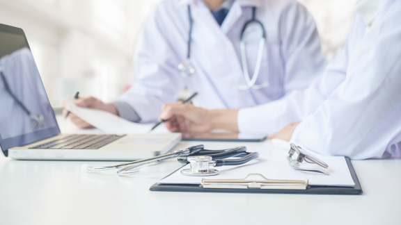 Ärzte besprechen Unterlagen am Laptop, mit Stethoskop auf dem Tisch als Symbol für das Gesundheitssystem