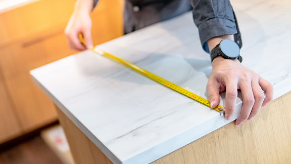 Hand misst eine weiße Arbeitsplatte mit Marmorstruktur für die Küchenmontage. Präzision beim Einbau.