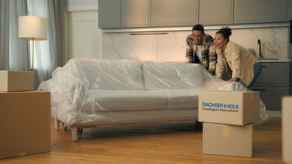 Zwei Personen schieben ein mit Schutzfolie verpacktes Sofa in einer Wohnung zwischen Umzugskartons