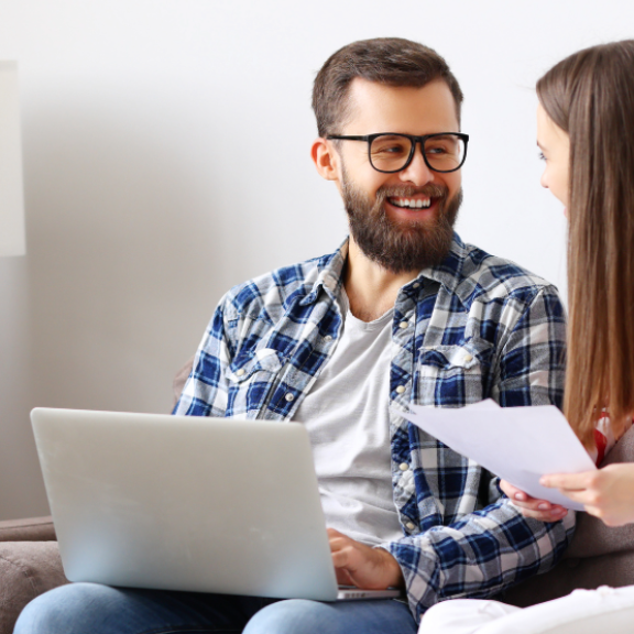 Glückliches Paar plant Umzug mit Laptop und Dokumenten, um Transportversicherung abzuschließen. Umzugskartons im Hintergrund.