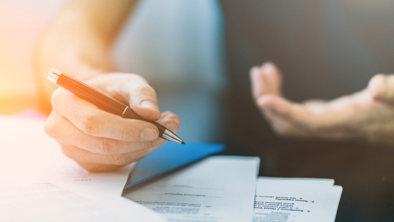 Hand unterschreibt Dokumente, möglicherweise für eine Transportversicherung. Im Hintergrund eine sprechende Person.